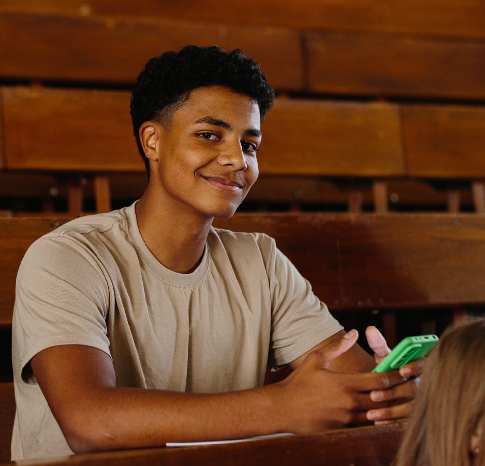 A young male students sits in a gymnasium and looks at the camera with a green phone in his hand.